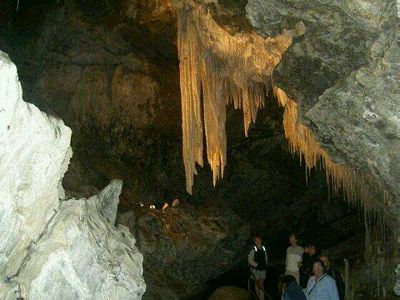 Veils of pale-gold stalactites hang from the ceiling of this cave. Chunky masses of white rock dominate the foreground.